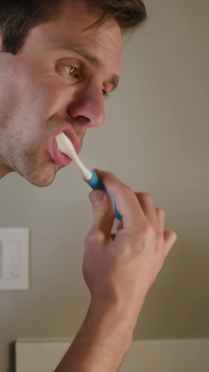 Vertical video: Leaning man in tan tee brushing teeth with power brush cleaning at sink, copy space