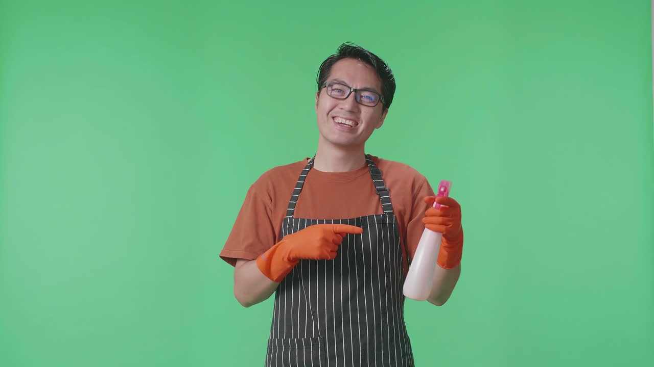초록색 스크린 스튜디오에 서 있는 동안 스프레이를 가리키는 아프론을 입은 미소 짓는 아시아 남성 집주인