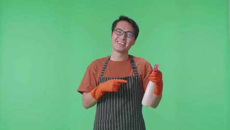 sonriente ama de casa masculina asiática con un delantal apuntando a un spray mientras está de pie en el estudio de pantalla verde