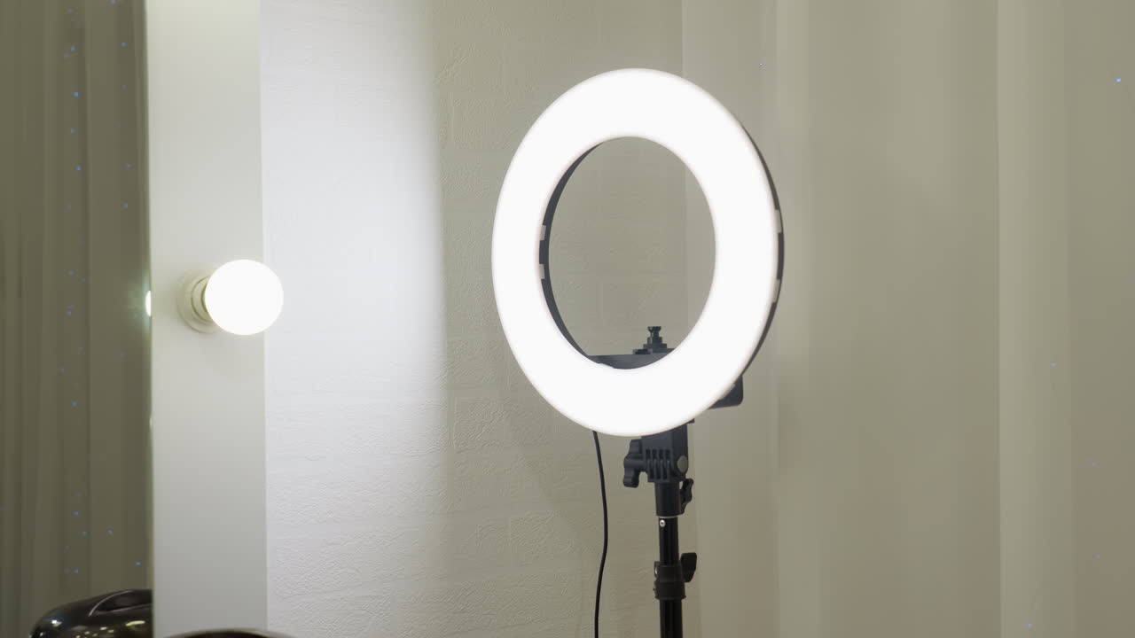 Bright ring light on stand positioned beside lit bulb and vertical mirror in salon setting, reflecting clean and minimalistic workspace with modern lighting tools and neutral curtain background