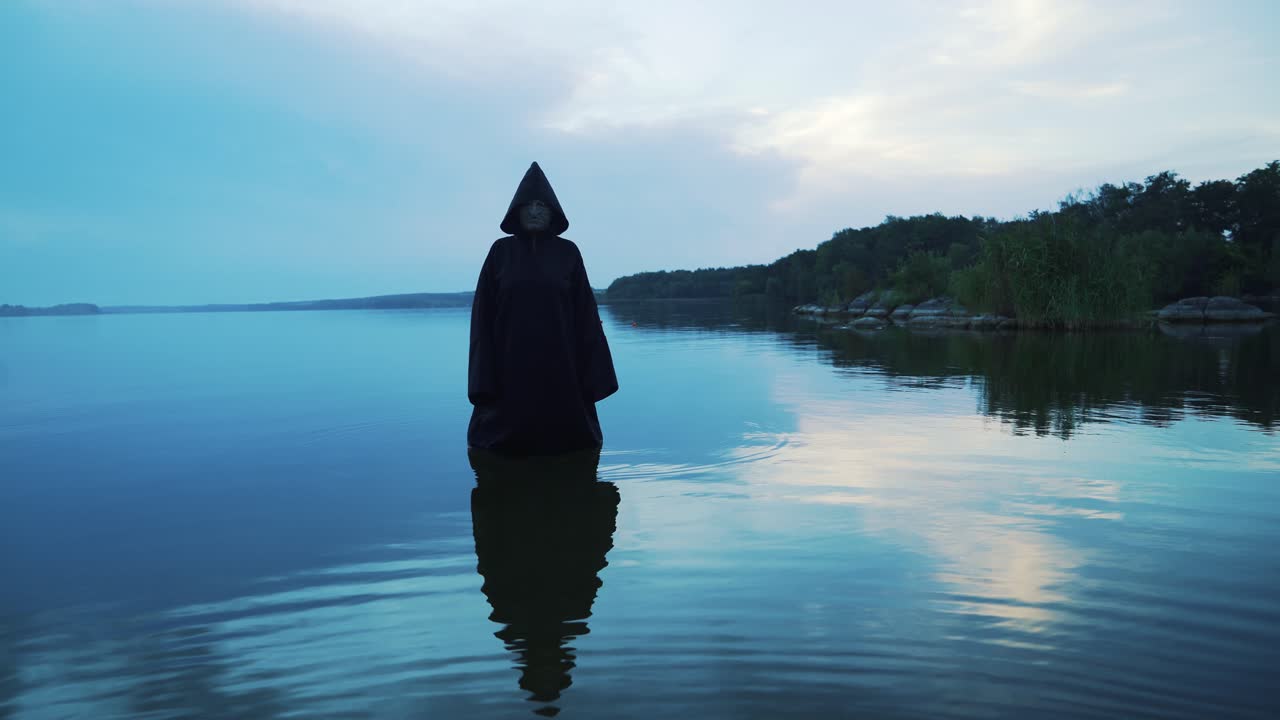 Evil witch in black cloak with hood in the river. Halloween costume.