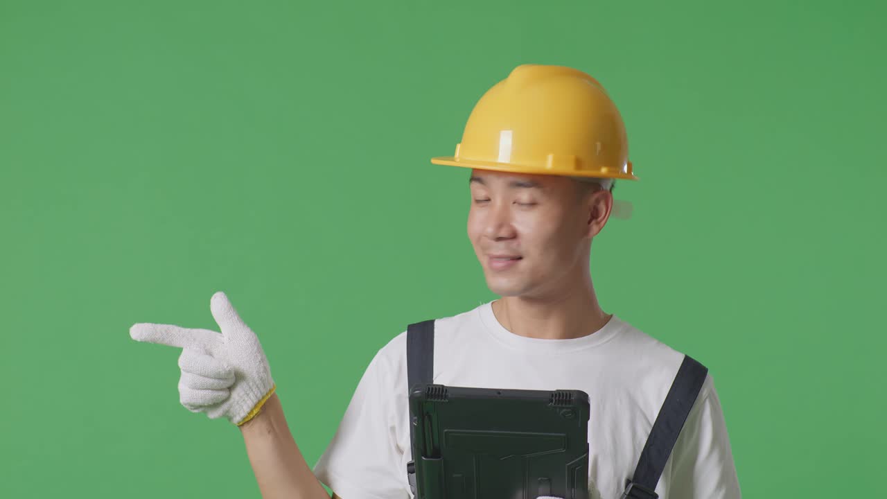 Close Up Of Asian Man Painter Wearing Safety Helmet Using A Tablet And Pointing To Side While Standing In The Green Screen Background Studio