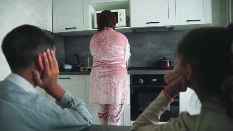 Boy and girl sit at kitchen table watching woman in pink robe prepare food at microwave, morning light filtering through window, creating intimate family moment in cozy modern home kitchen interior