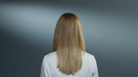 Parte-Trasera-De-La-Joven-Y-Atractiva-Doctora-Rubia-Con-La-Bata-Médica-Blanca-Girando-Hacia-La-Cámara-Y-Sonriendo-Alegremente-En-El-Fondo-De-La-Pared-Gris