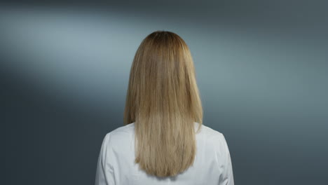 흰색 의료 가운에 금발의 젊은 매력적인 여성 의사의 후면은 카메라를 돌리고 회색 벽 배경에 유쾌하게 웃고