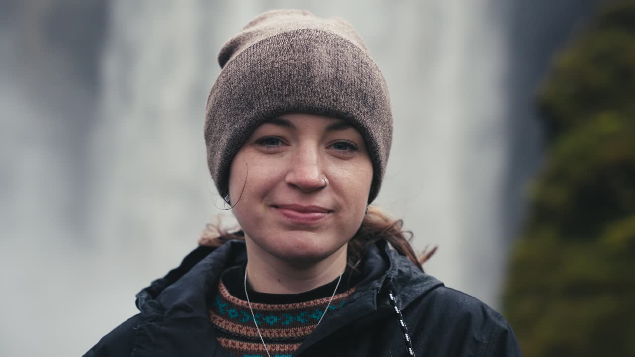 귀고 아름다운 하이킹 소녀가 화려한 폭포 앞에서 카메라를 향해 미소 짓고 있다.