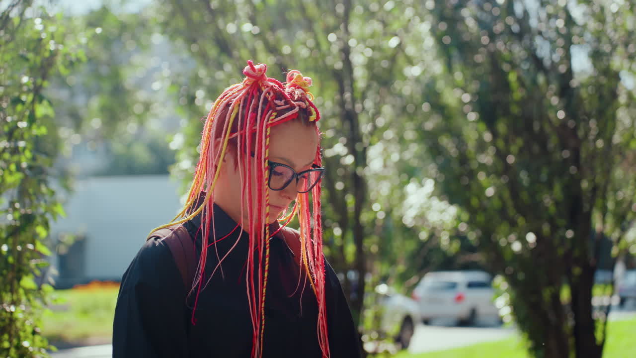 Mujer caucásica abatida con trenzas rosas cerca de árboles al borde de la carretera y coches aparcados, gafas, pausa tranquila durante un descanso, la suave luz del sol y el bokeh crean un retrato melancólico al borde de la carretera lleno de reflexión