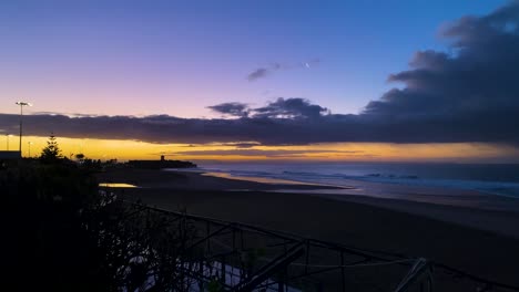 panoramic-shot-revealing-some-waves-breaking-on-the-beach-towards-the-sunrise-with-the-moon-in-the-background-in-Carcavelos,-Lisbon,-Portugal