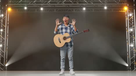 cuerpo lleno de adolescente asiático con guitarra mostrando gestos manos palma, diciendo wow, y sonriendo en el escenario