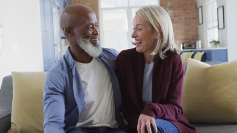 portrait of happy senior diverse couple in living room sitting on sofa, embracing and smiling