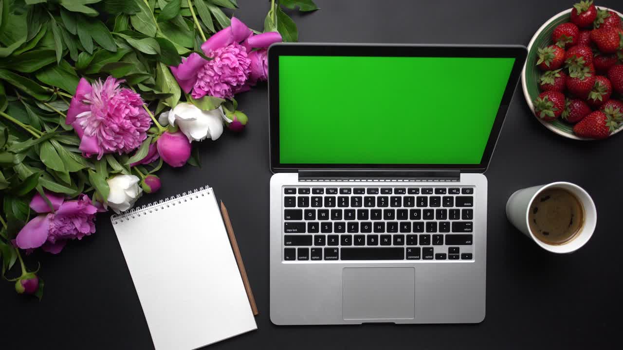 Laptop computer device with green screen, notebook, cup of coffe and beautiful bouquet of pink and white peony flowers on the black background. Table top view. Chroma key