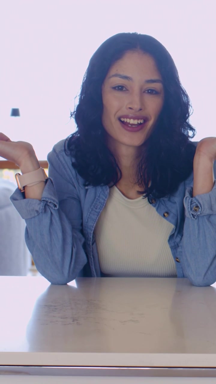 Video verticale: Donna sorridente in jeans appoggiata a un tavolo vicino alla finestra, che spiega gesticolando con i palmi delle mani.