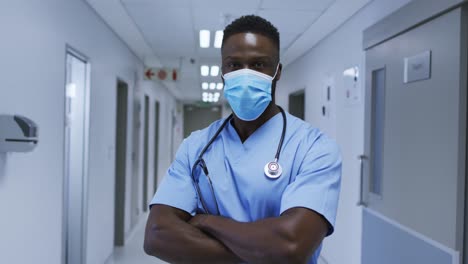 Portrait-of-african-american-male-doctor-wearing-face-mask-and-scrubs-standing-in-hospital-corridor