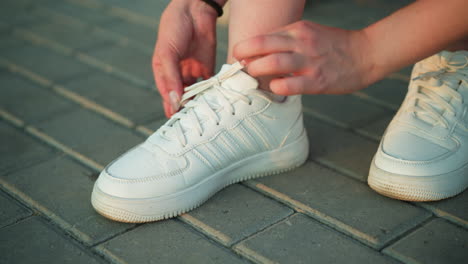 close-up of person tying shoelaces of white sneakers on interlocked pavement path, focusing on hands firmly adjusting laces for a secure fit