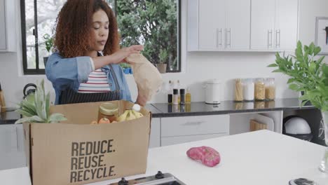 Happy-biracial-woman-unpacking-groceries-from-recycled-box-in-kitchen,-copy-space,-slow-motion
