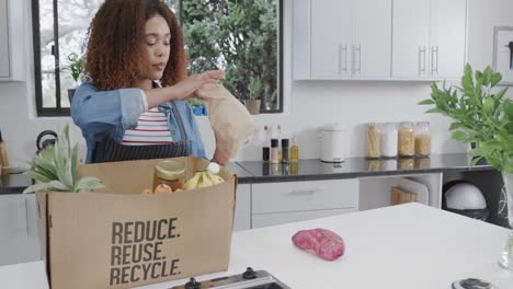 una mujer biracial feliz desempaquetando comestibles de una caja reciclada en la cocina, espacio de copia, cámara lenta.