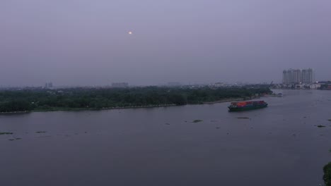 Barco-Contenedor-Que-Transporta-Carga-En-El-Río-Saigón,-Vietnam-Al-Atardecer-Con-Vista-Al-Agua-Y-La-Luna-Llena