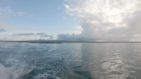 a boat is moving away from an island of the galapagos islands. while it moves it creates small waves on the blue ocean. on the back a blue sky and clouds