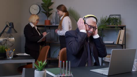 joyful senior businessman working on portable laptop sitting at desk, listening music and dancing