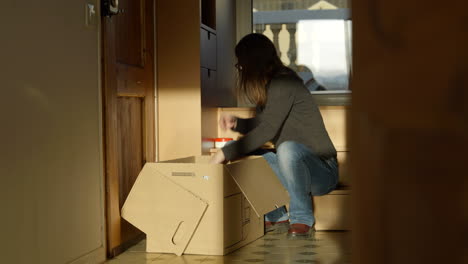 mujer sentada en la sala de estar iluminada por el sol empacando cajas de cartón preparándose para una nueva mudanza