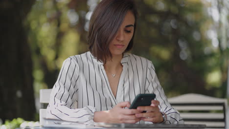 young hispanic latin ethnic woman sitting in a summer cafe holding phone looking at screen watching social media video content movie or stream online in mobile app laughing using smart phone