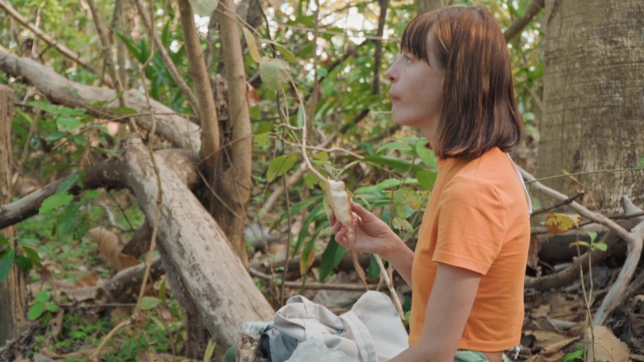 Primer plano de una mujer examinando y comiendo un trozo de pan en un entorno forestal con denso follaje verde y troncos de árboles. La mujer sostiene comida parcialmente comida mientras está sentada en el suelo junto a una bolsa