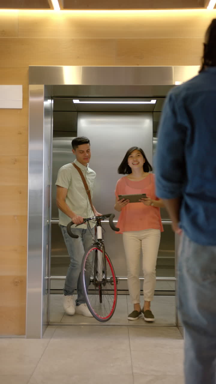 Vertical video: Man with bicycle and woman holding tablet exiting elevator, greeting colleague