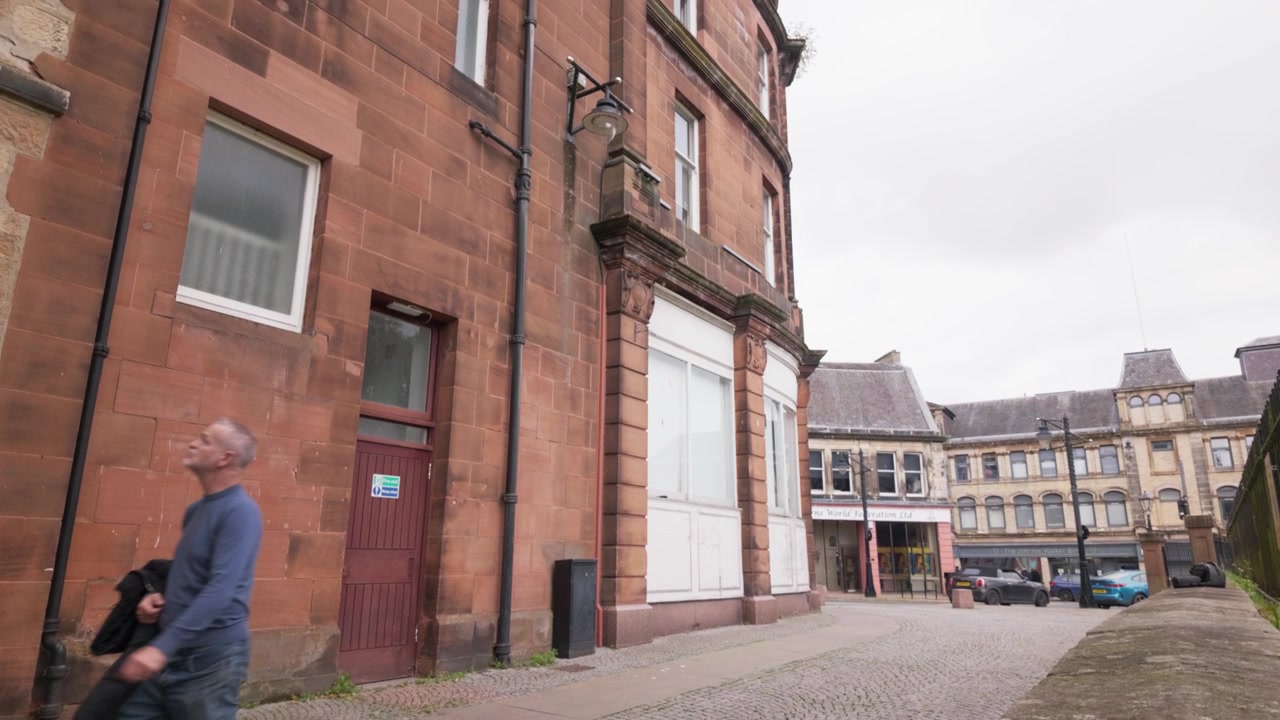 Eine Person geht durch eine ruhige Straße in Kilmarnock, Schottland, vorbei an traditionellen Steingebäuden und historischer Architektur in einer ruhigen städtischen Umgebung, Stativ-Tracking-Aufnahme