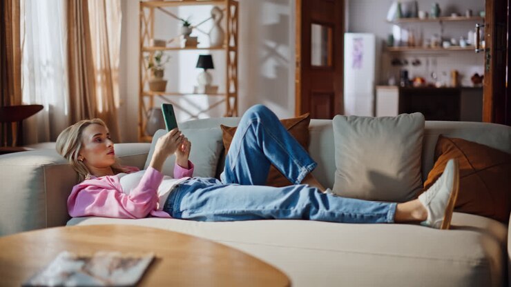 Online pige ligger i sofaen og sms'er på sin mobiltelefon. Blond kvinde slapper af på sofaen derhjemme