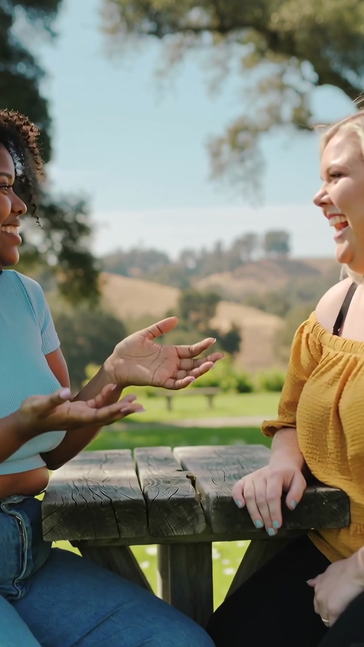 Vertical video: Speaking left friend in blue gesturing at table, right friend laughing in mustard