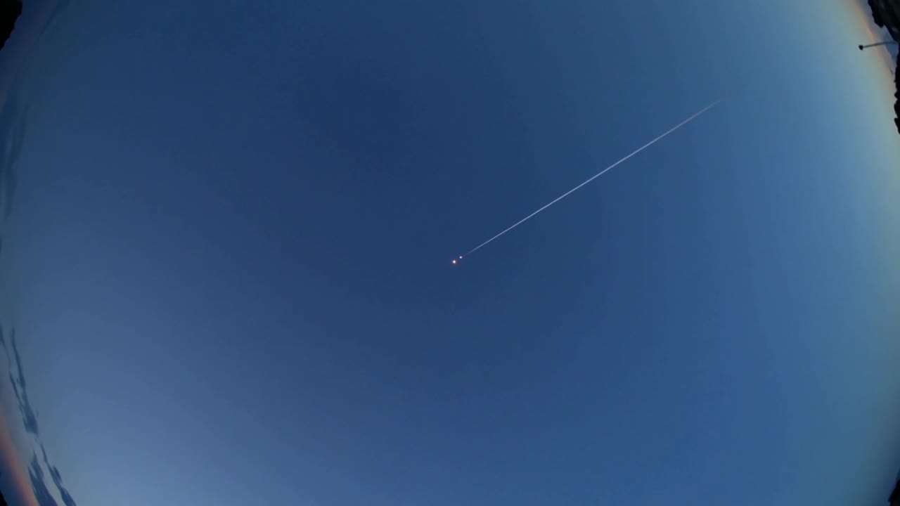Aereo con due luci, visibile in alto a destra, diretto a destra verso l'orizzonte nel cielo crepuscolare con scia di condensazione, effetto fisheye.