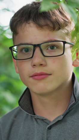 primer plano de un niño con labios rosados, con gafas y una camiseta gris, parpadeando mientras mira directamente a la cámara, está rodeado de exuberante vegetación con árboles borrosos en el fondo