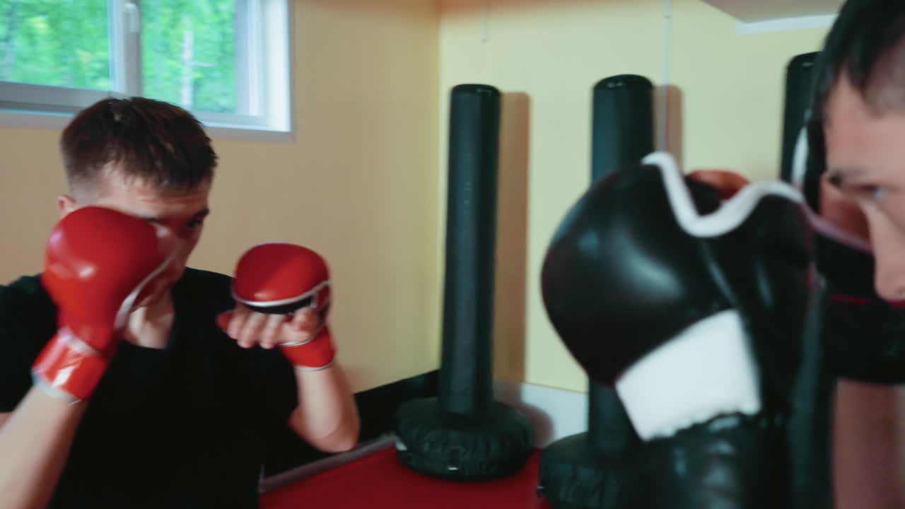 체육관에서 스파링하는 격투가들, 빨간 글러브를 낀 남자가 얼굴을 가리고 있고 검은 글러브를 낀 상대는 앞으로 나아가고 있어요. 둘 다 창문에서 들어오는 자연광 아래에서 강렬한 전투 훈련에 집중하고 있어요.