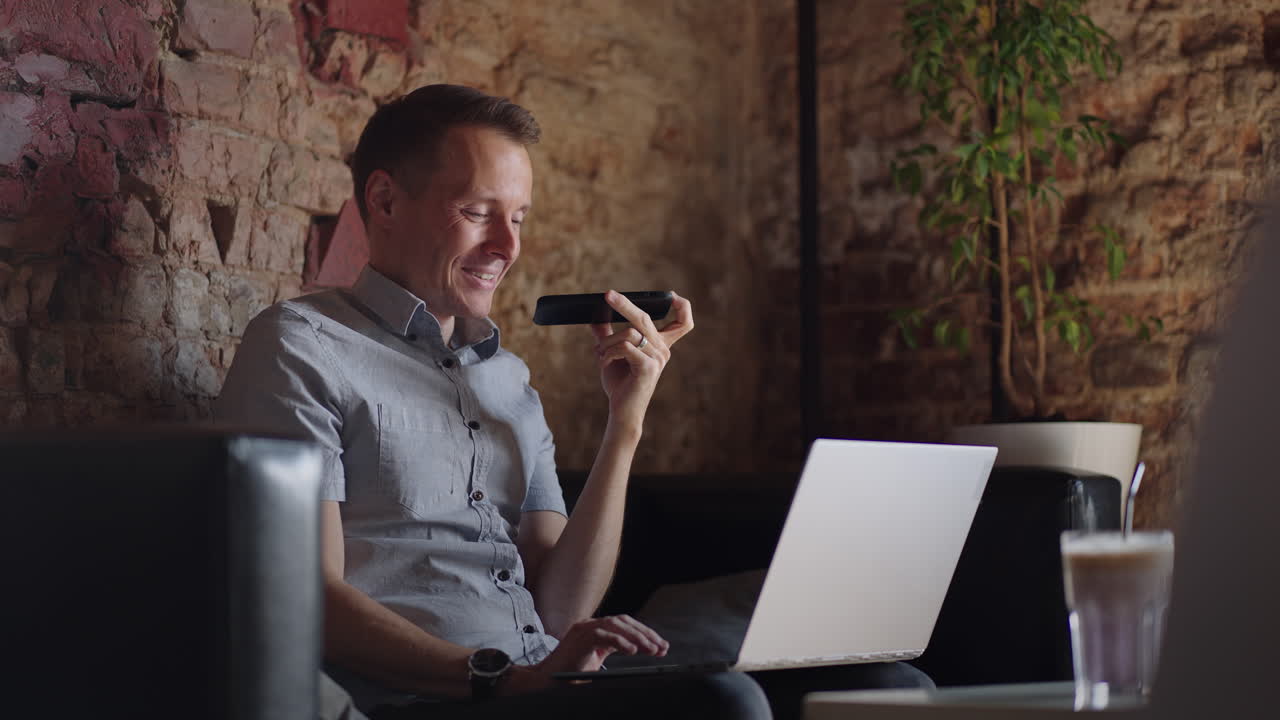 Smiling young man recording voice recognition message on speakerphone. Businessman using virtual assistant app sets reminder on smart phone at home office. Digital ai mobile assistance tech concept