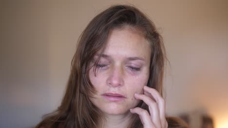 A-close-up-portrait-shot-of-a-young-woman-with-wounds-and-bruises,-looks-and-stares-directly-into-the-camera-while-touching-her-sore-cheek-and-starts-crying