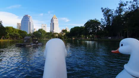 POV-Aufnahmen:-Köpfe-Eines-Schwanenpaddelboots-Auf-Dem-See-Mit-Wolkenkratzern-Im-Hintergrund-Im-Lumphini-Park,-Bangkok,-Thailand