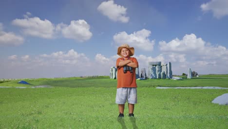 asian boy with a hat and binoculars crossing arms and smiling to camera while traveling in stonehenge. boy researcher examines something, travel tourism adventure, full body