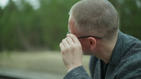 una vista de cerca de un hombre ajustando sus gafas mientras mira hacia el lado, capturado al aire libre con un fondo verde y borroso, el hombre lleva una chaqueta gris