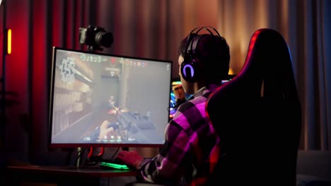 vista trasera de streamer chico asiático gritando gol y bailando celebrando el juego ganador a través de la red en la computadora personal. videojuego de transmisión en vivo, escritorio iluminado por luz de tira de led rgb