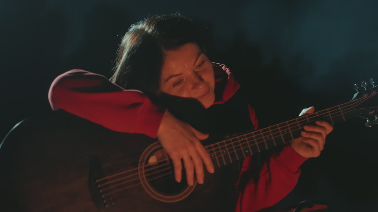 Donna asiatica in campeggio, chitarra, falò di notte; musicista camper solitaria che strimpella una melodia acustica intensa, accogliente bagliore di brace e fumo che si disperde, primo piano spontaneo di una suonatrice sorridente appoggiata allo strumento, intima.