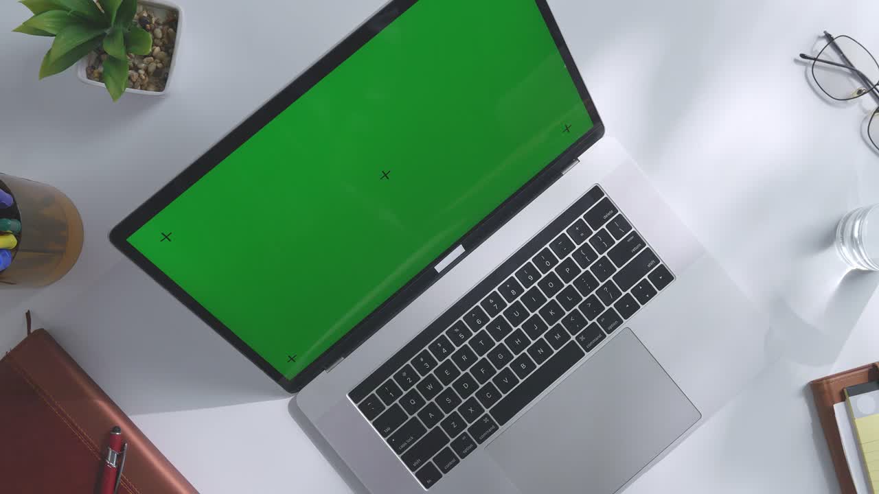 vista de arriba hacia abajo de una computadora portátil con pantalla verde simulada pantalla chromakey en un escritorio de oficina de madera al lado de la libreta con bolígrafos, gafas y un vaso de agua.