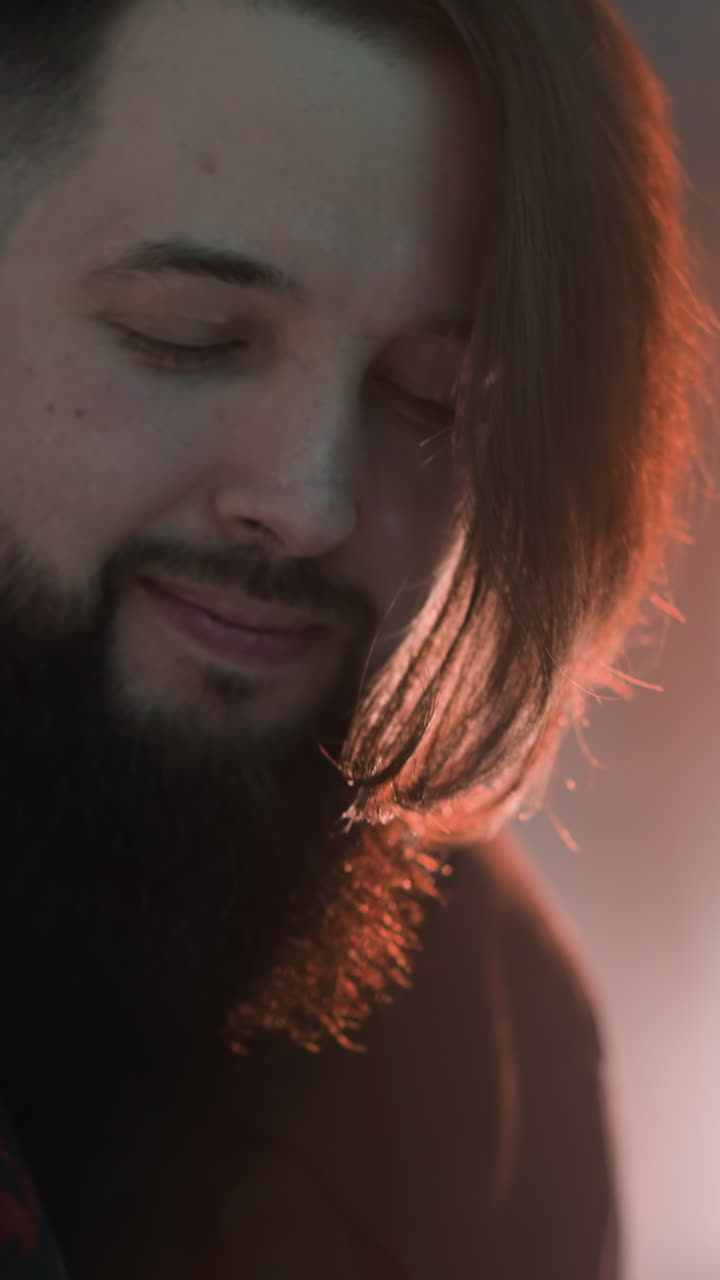 guitarrista barbudo con una camisa negra tocando la guitarra en una habitación suavemente iluminada y borrosa. la iluminación ambiental destaca la expresión enfocada del músico