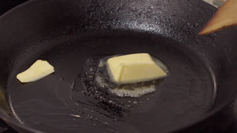 close up: pat of butter melts in hot cast iron pan, wood spatula stirs