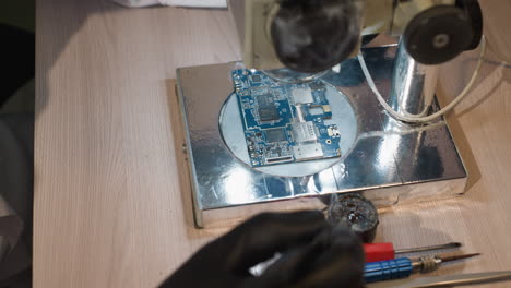una vista de cerca de un banco de trabajo de reparación de electrónica con un técnico soldando una placa de circuito bajo un microscopio, varias herramientas están esparcidas alrededor