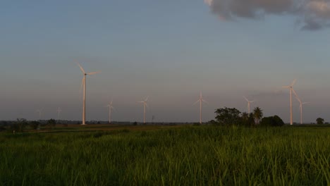 Windkraftanlagen-Schießen-Aus-Einem-Ackerland,-Während-Die-Sonne-Untergeht,-Während-Lastwagen-Vorbeifahren-Und-Staubwolken-Erzeugen,-Saubere-Alternative-Energie-In-Thailand-Und-Auf-Dem-Südostasiatischen-Festland