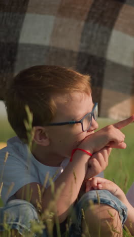 a woman in a blue gown, her face not visible, holds a plaid scarf as the wind gently blows it in a grassy field. beneath the scarf, two happy children in white shirts point at their surroundings