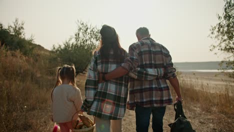 Rear-view-of-a-happy-couple,-a-brunette-girl-in-a-Green-checkered-shirt-and-a-Green-hat,-walks-and-hugs-with-her-husband-with-gray-hair-and-their-little-daughter-is-walking-next-to-them-on-a-picnic-along-the-road-with-dry-grass