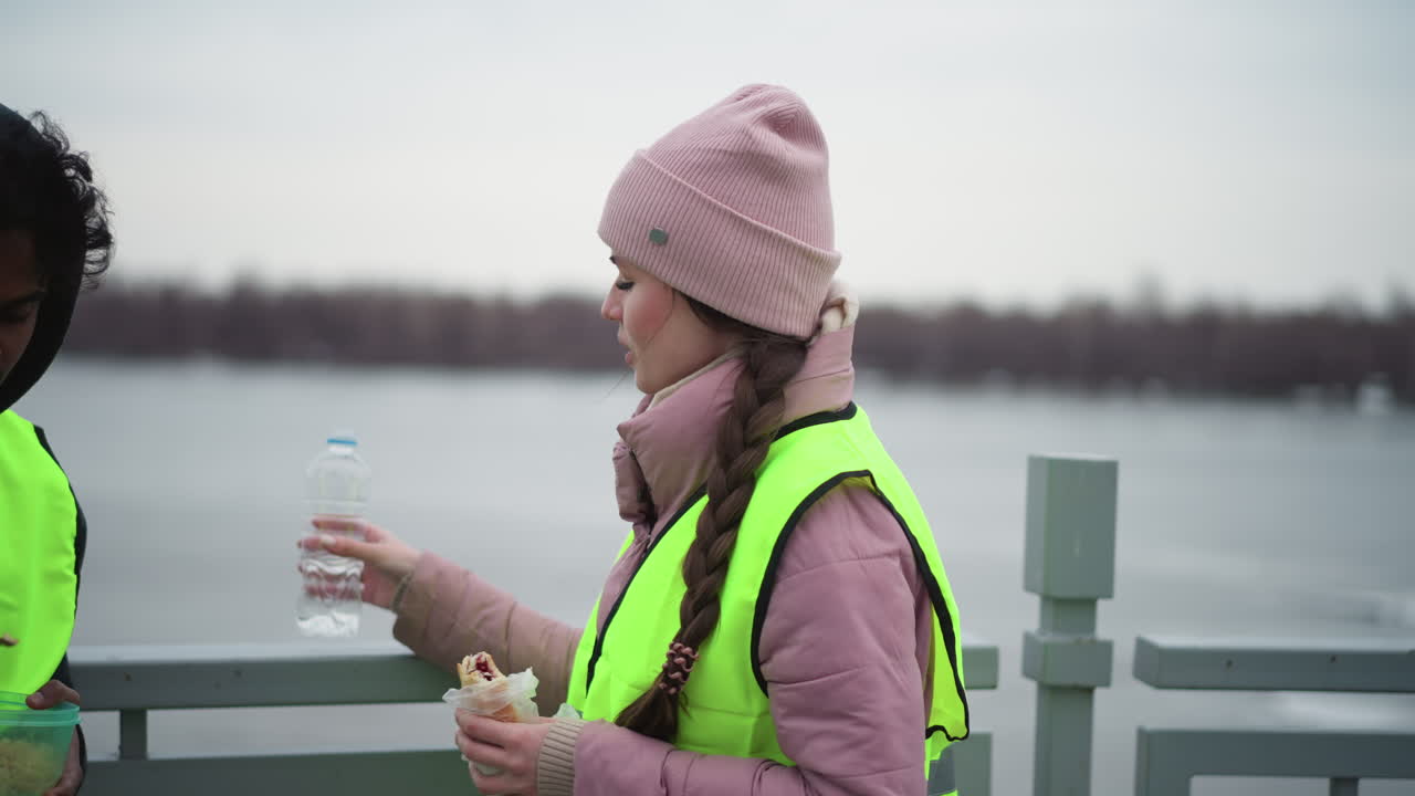 Smiling woman in pink jacket and beanie wearing neon safety vest holding snack and reaching for water bottle outdoors near river on cold overcast day, depicting concept of teamwork, outdoor work