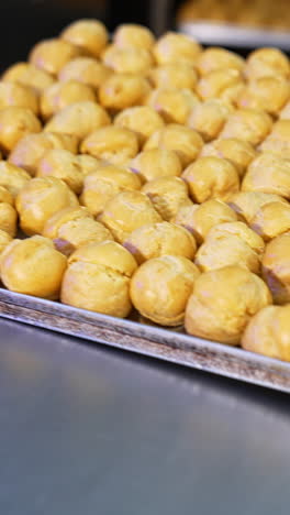 freshly baked profitroles being taken off the oven. factory worker pulls the baking sheet from oven and puts it on the table. close up. vertical video
