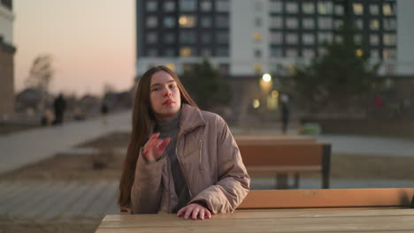 a young woman dressed in a peach jacket and black trousers walks towards a bench in an urban park. after sitting down, she places her hand on the table and gazes off with an unhappy expression